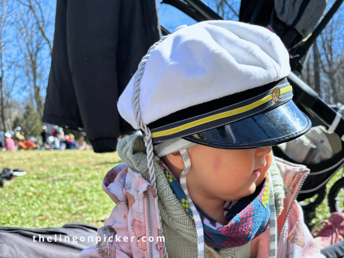 Toddler wearing a white graduation cap at a family picnic, celebrating May Day in Finland during spring, with green grass and a festive atmosphere.